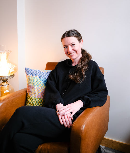 A woman with brown hair tied back sits smiling in a brown leather armchair, wearing a black hoodie and black pants. A colorful patterned throw pillow rests beside her, and a lit table lamp is visible against a plain light gray wall.